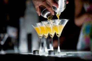 Photograph of human hands holding two cocktail shakers and pouring orange color cocktail into two martini glasses